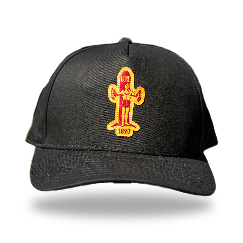 Black curved-brim cap with red and gold felt patch of Duke Kahanamoku standing with surfboard and 1890